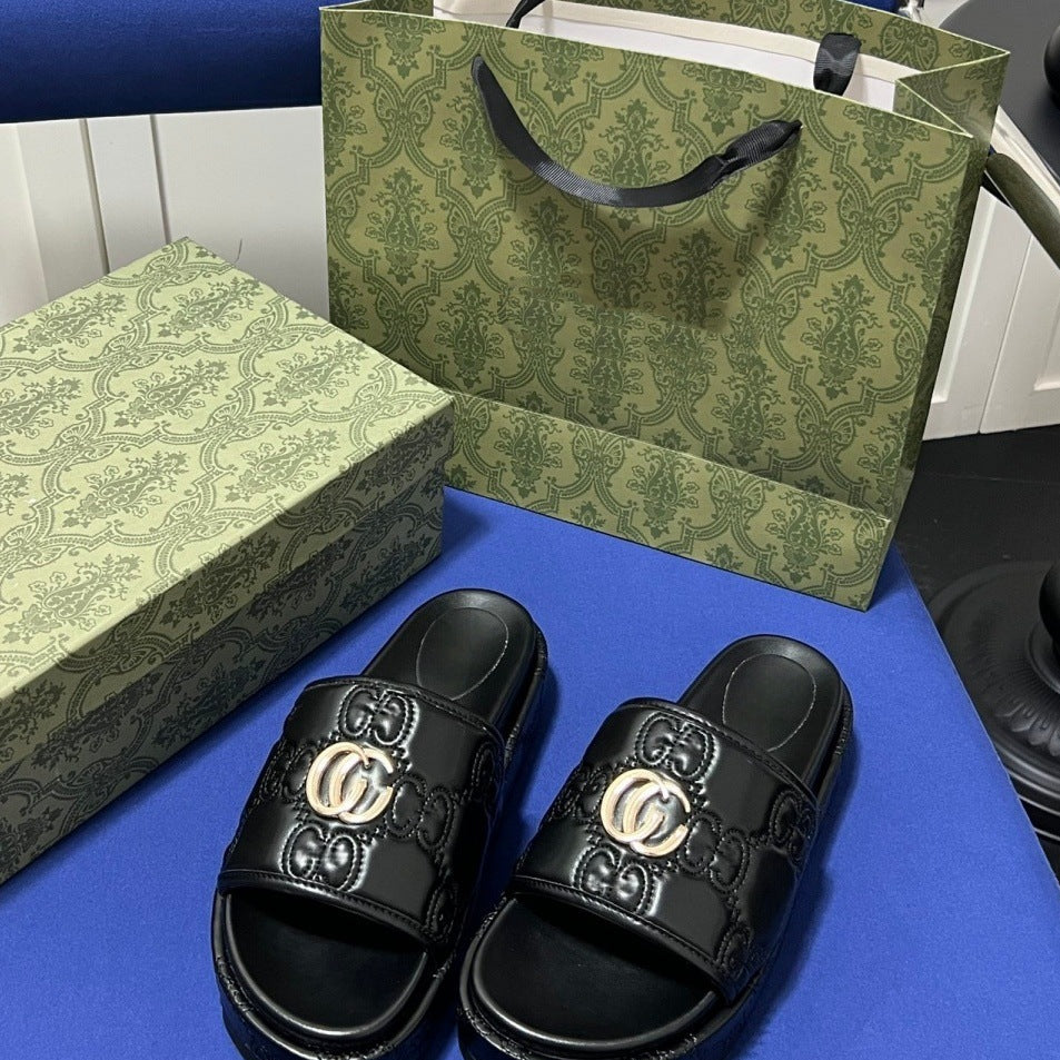 A pair of black Gucci slide sandals with embossed GG monogram details and a gold-tone interlocking G logo on the strap, displayed with a matching green patterned shoebox and shopping bag on a blue surface.