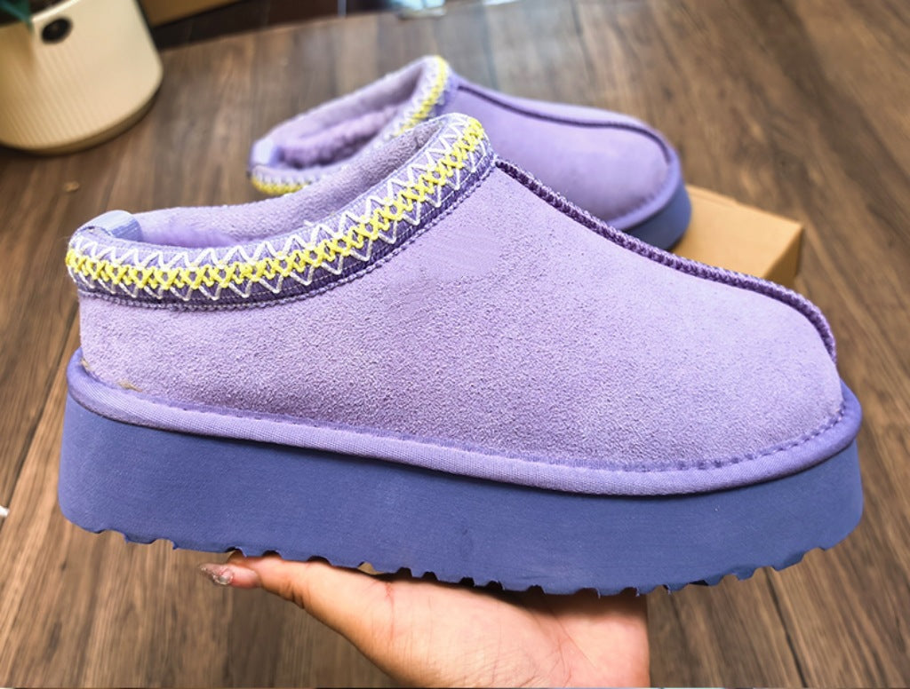 A pair of purple slip-on mule slippers with a thick blue sole and yellow-and-white zigzag trim, shown with one being held by a hand over a wooden floor.