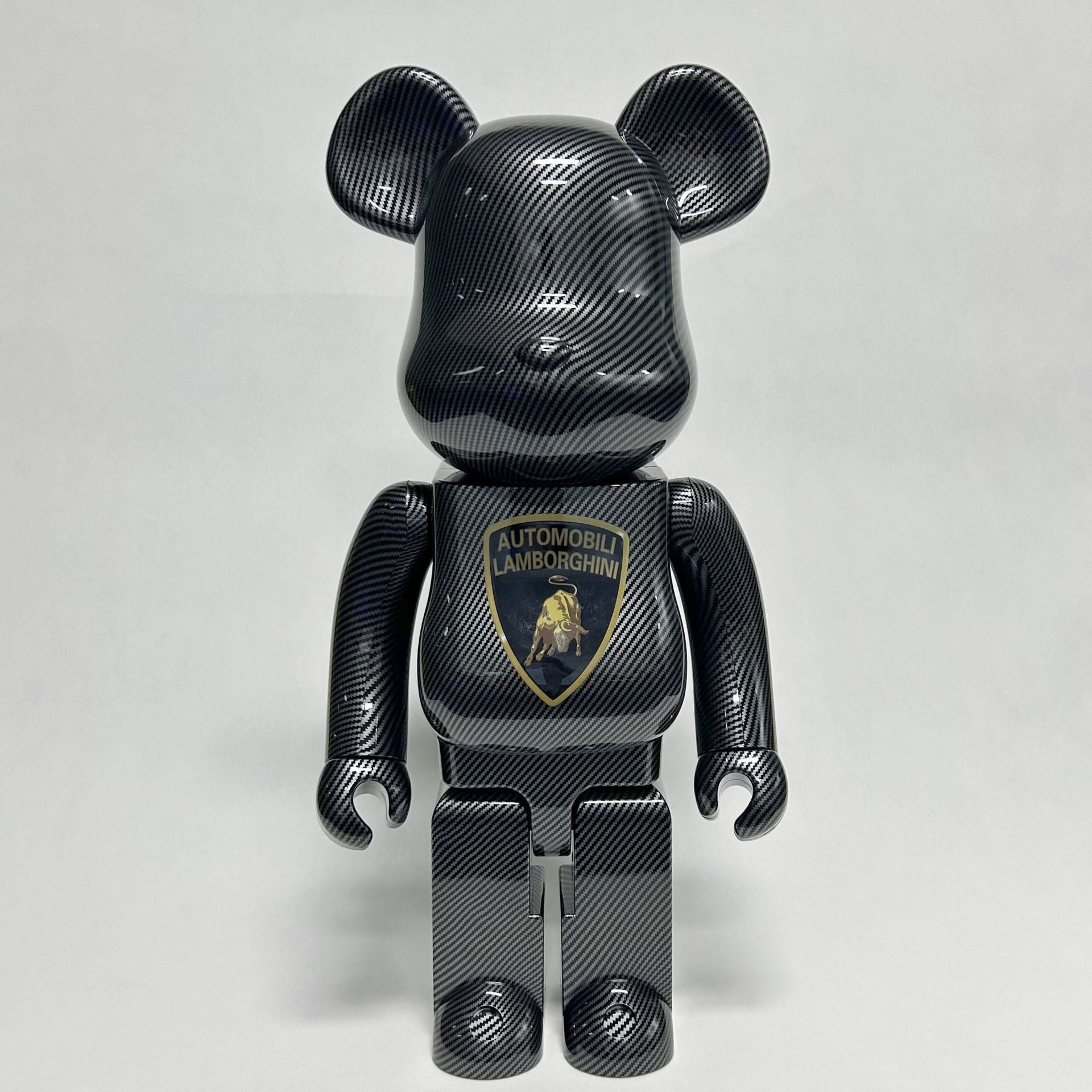 A black carbon-fiber-patterned bear-shaped collectible figure with a Lamborghini chest badge, photographed against a light gray studio background.