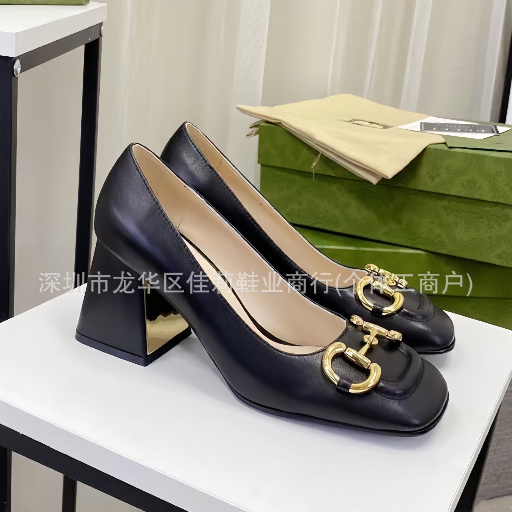 A pair of women's black leather block-heel pumps with a gold horsebit detail on the toe, displayed on a white table with a green shoebox in the background.