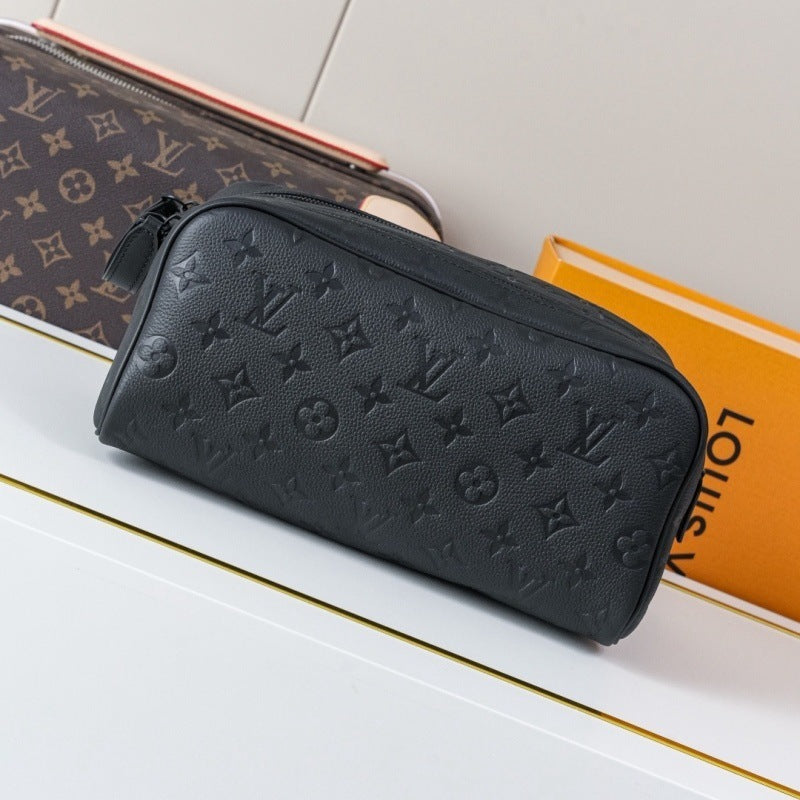 A black textured leather Louis Vuitton toiletry bag with embossed monogram pattern, displayed on a white surface next to a Louis Vuitton monogram pouch and an orange Louis Vuitton box.