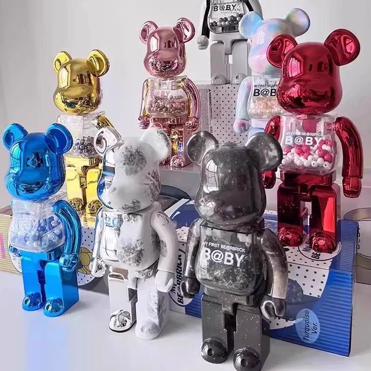 A group of translucent bear-shaped collectible figures with metallic and distressed finishes, including blue, gold, pink, red, white, and dark gray/black versions, arranged on a white surface with a branded display box behind them.