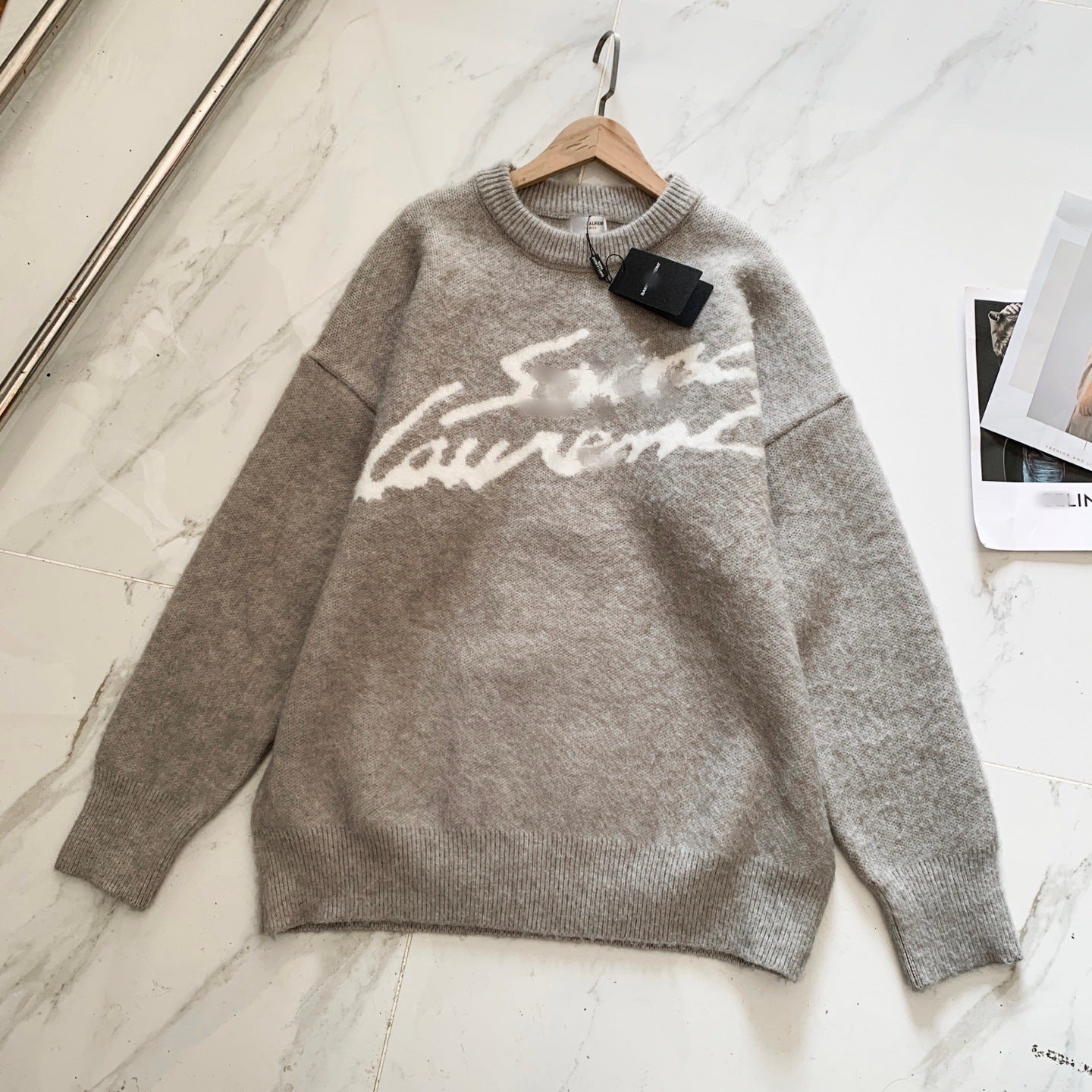 A light gray knit sweater with a white 'Saint Laurent' script across the chest, hanging on a wooden hanger against a white marble-patterned background.