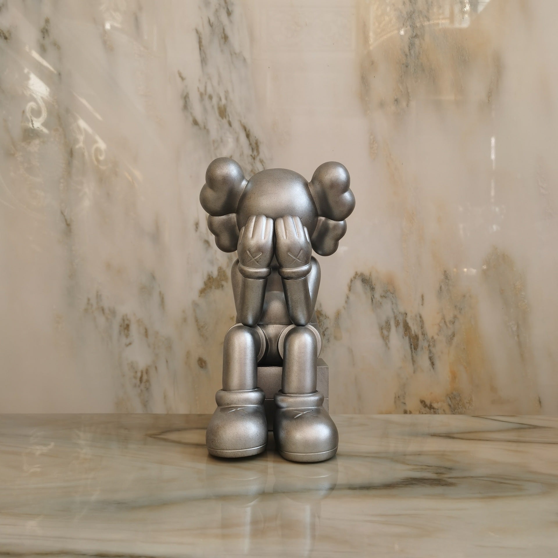 A metallic silver KAWS-style figurine seated on a marble surface against a marble-patterned background.