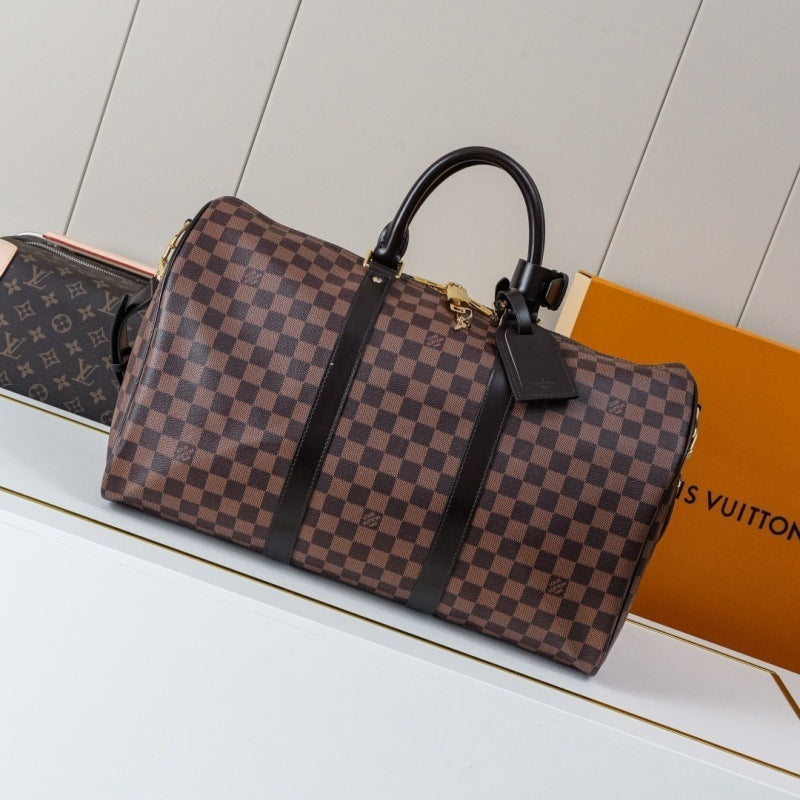 A Louis Vuitton monogram duffel bag in brown and beige checkered canvas with black leather handles and straps, displayed on a white surface next to an orange Louis Vuitton box.
