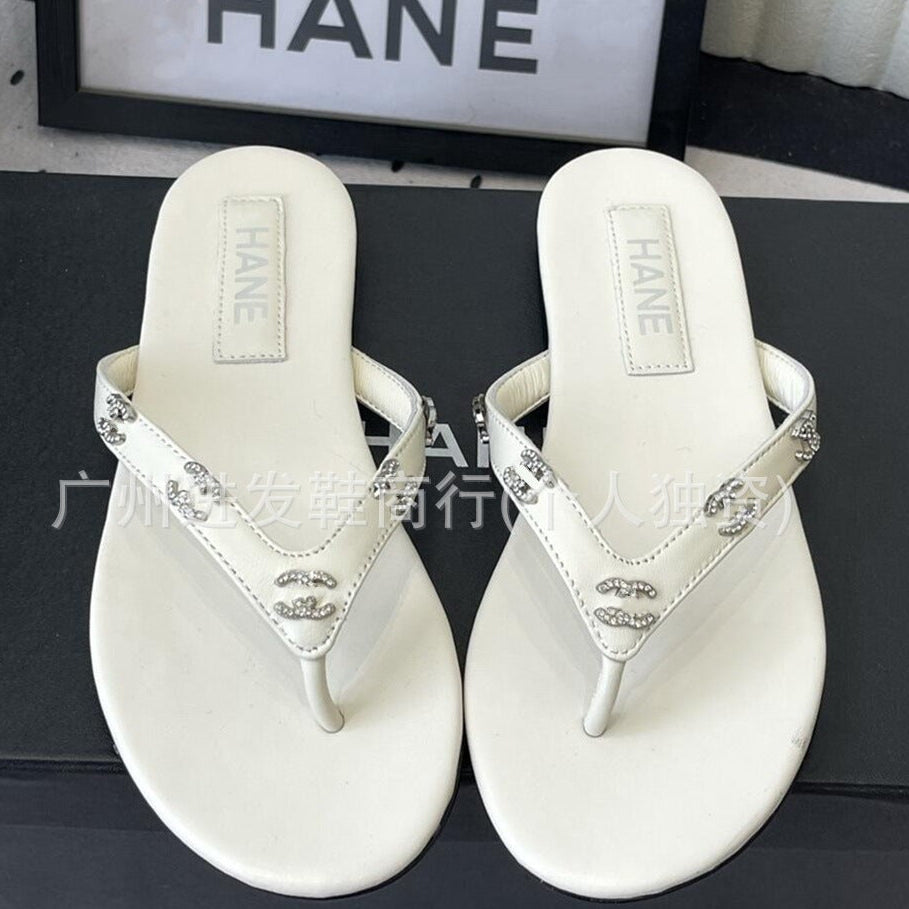 A pair of white leather thong sandals with silver-tone rhinestone straps and a double-C logo detail, displayed on a black surface with a Chanel-branded box in the background.