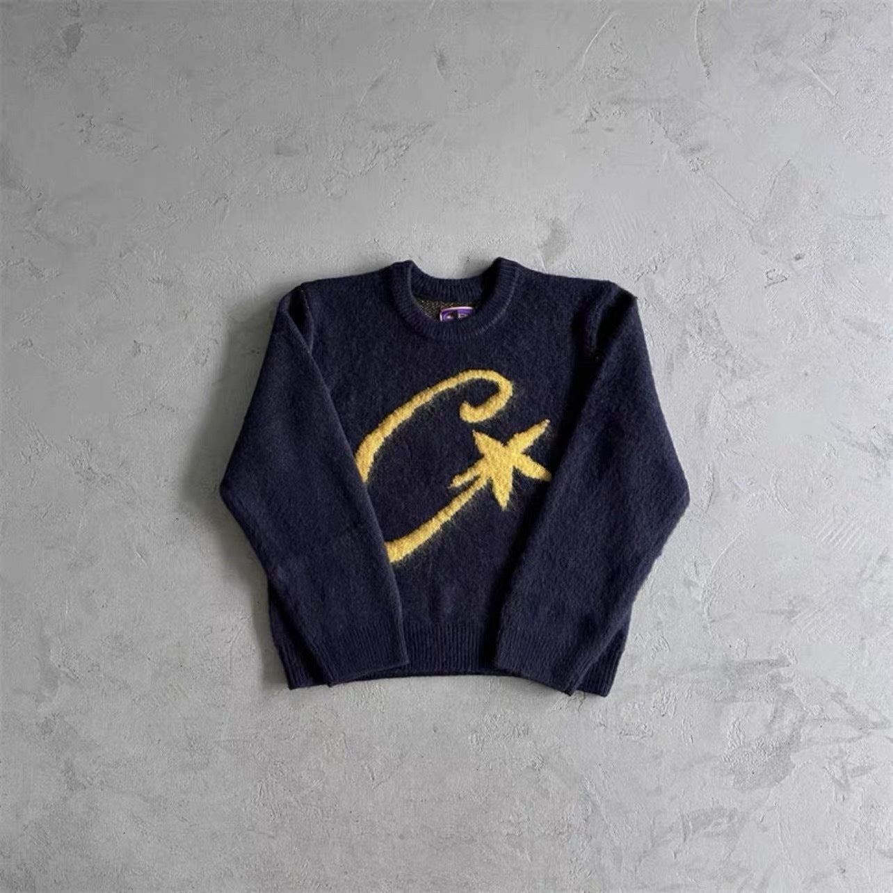 A dark navy blue knit sweater with a large yellow embroidered cursive 'C' and star motif on the front, laid flat on a textured gray concrete surface.