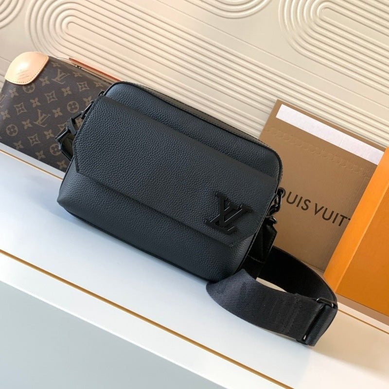 A black textured leather crossbody bag with a black adjustable strap and a prominent black LV logo on the front, displayed on a white surface with Louis Vuitton packaging in the background.