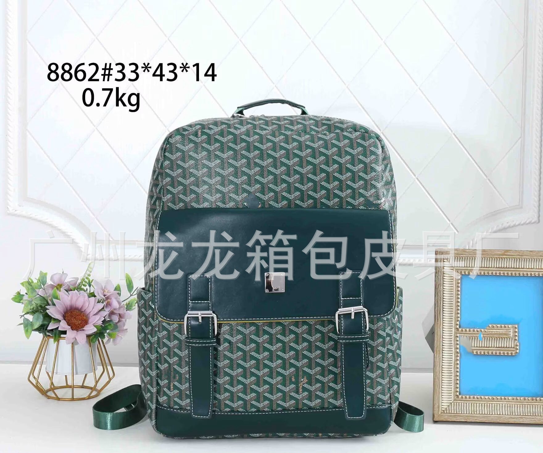 A Goyardine backpack in green and beige with dark green leather trim, displayed on a white surface with decorative props including a small flower vase and a picture frame. Text overlays show the model number and dimensions.