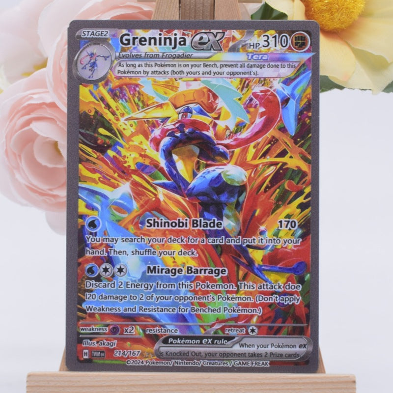 A Greninja ex Evoker Pokémon trading card displayed upright on a small wooden stand, with soft pink flowers blurred in the background.