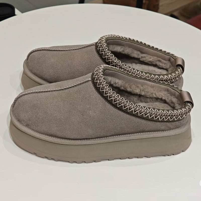 A pair of taupe suede slip-on slippers with a plush lining and decorative zigzag stitching around the collar, resting on a white round table.
