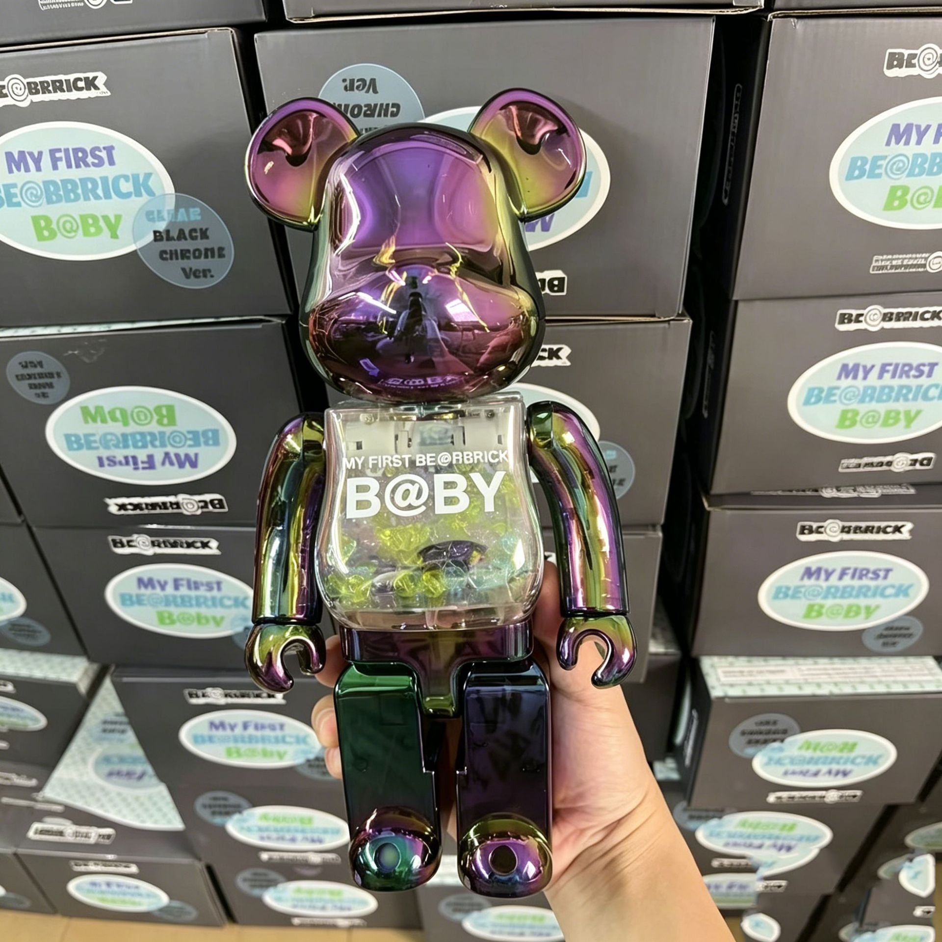 A hand holds a metallic iridescent purple and green Bearbrick B@by figure in front of stacked black boxes labeled BE BRICK and MY FIRST BE BRICK B@BY.
