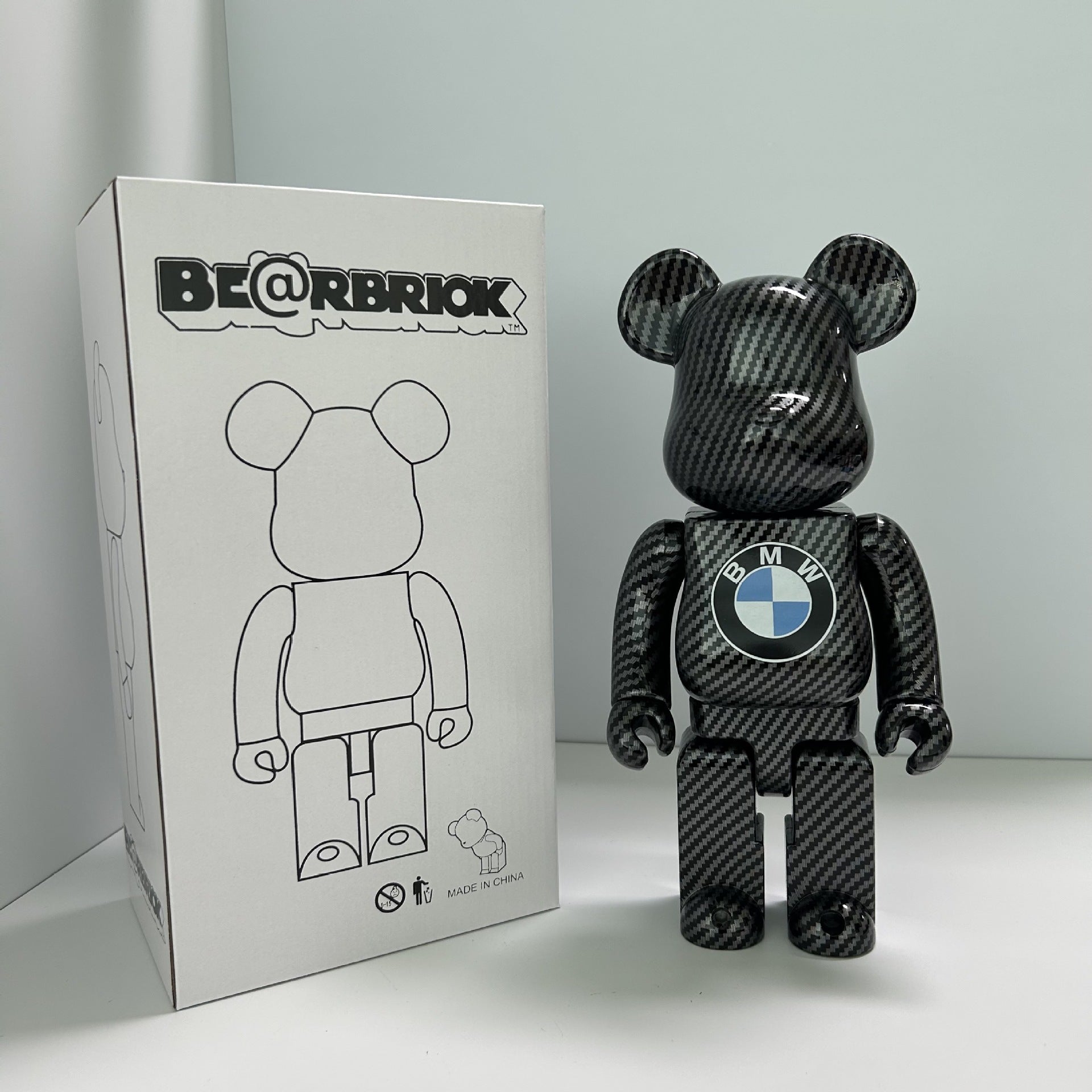 A Bearbrick x BMW collectible figure made of glossy black carbon fiber, standing next to its white packaging box on a white surface against a light gray background.