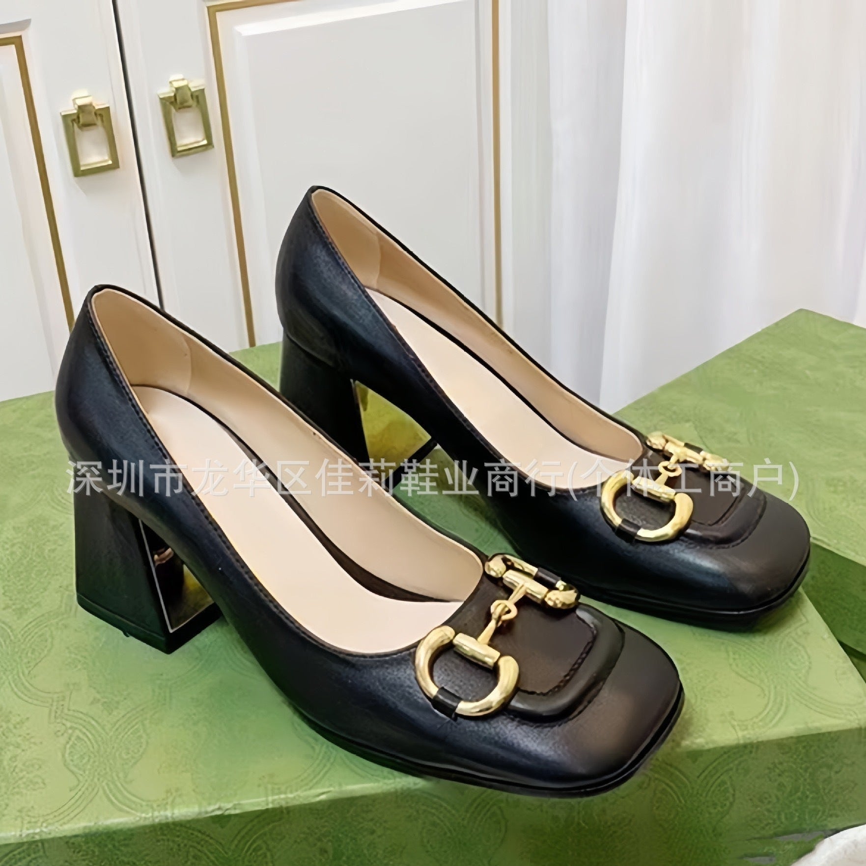 A pair of black leather high-heeled pumps with square toes and gold horsebit hardware, displayed on a green shoebox in front of white paneled doors.