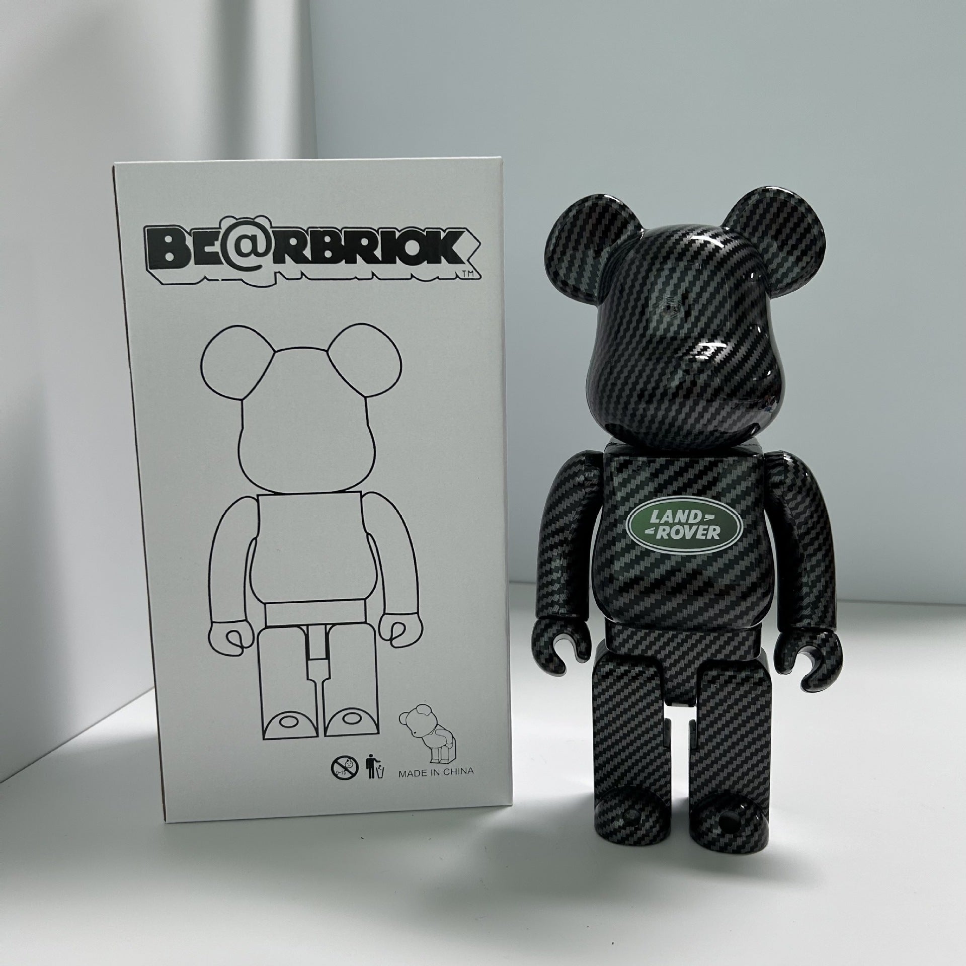 A Bearbrick collectible figure with a black carbon fiber pattern, standing next to its white packaging box. The figure features a Land Rover logo on its chest.