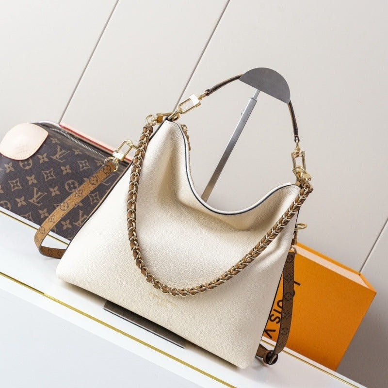 A cream-colored pebbled leather hobo handbag with gold-tone chain and strap hardware, displayed on a white shelf next to a Louis Vuitton monogram pouch and an orange box.