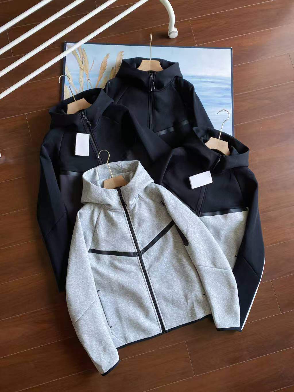 Three athletic zip-up jackets laid flat on a wooden floor: two black hooded jackets in the back and one heather gray zip-up hoodie in the front with black trim. No person or real environment beyond the floor backdrop is the focus, so this is Product/Packaging rather than Lifestyle.