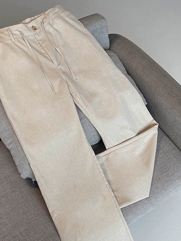 A pair of light beige straight-leg pants laid flat on a gray upholstered chair, with a small white printed brand mark near the lower right leg.
