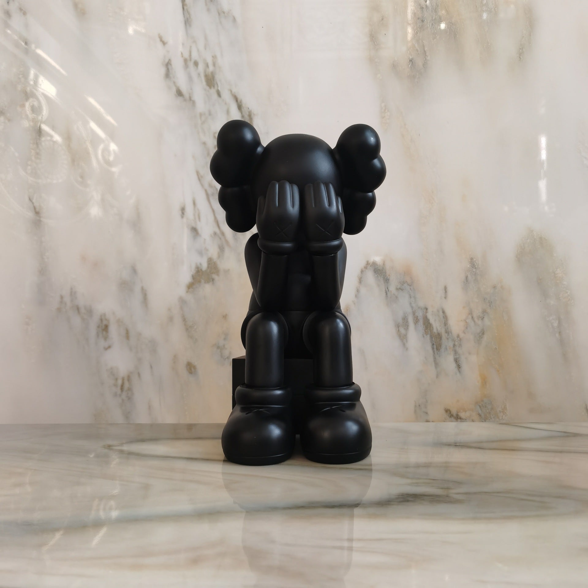 A matte black KAWS-style figurine seated with hands over its face, displayed on a polished marble surface against a matching marble wall.