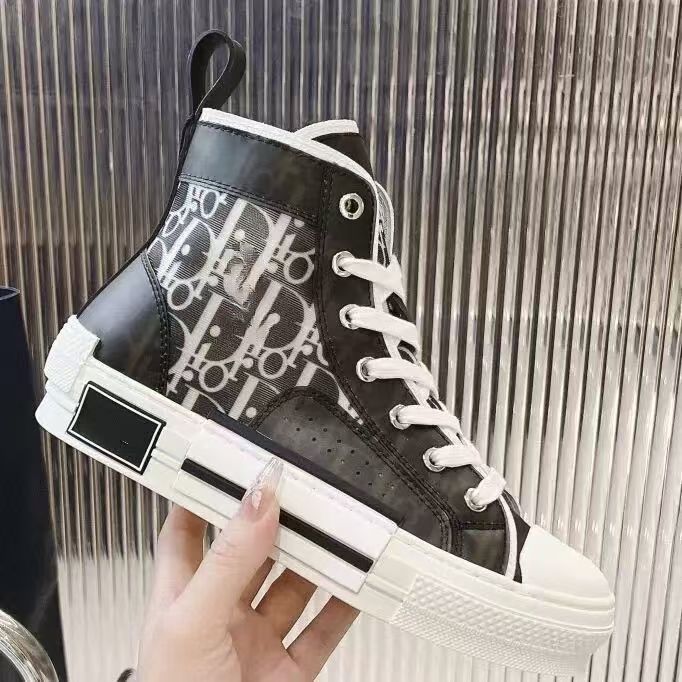 A high-top sneaker with a glossy black upper featuring a white Dior monogram pattern, white laces, and a chunky white sole, held by a hand against a metallic slatted background.