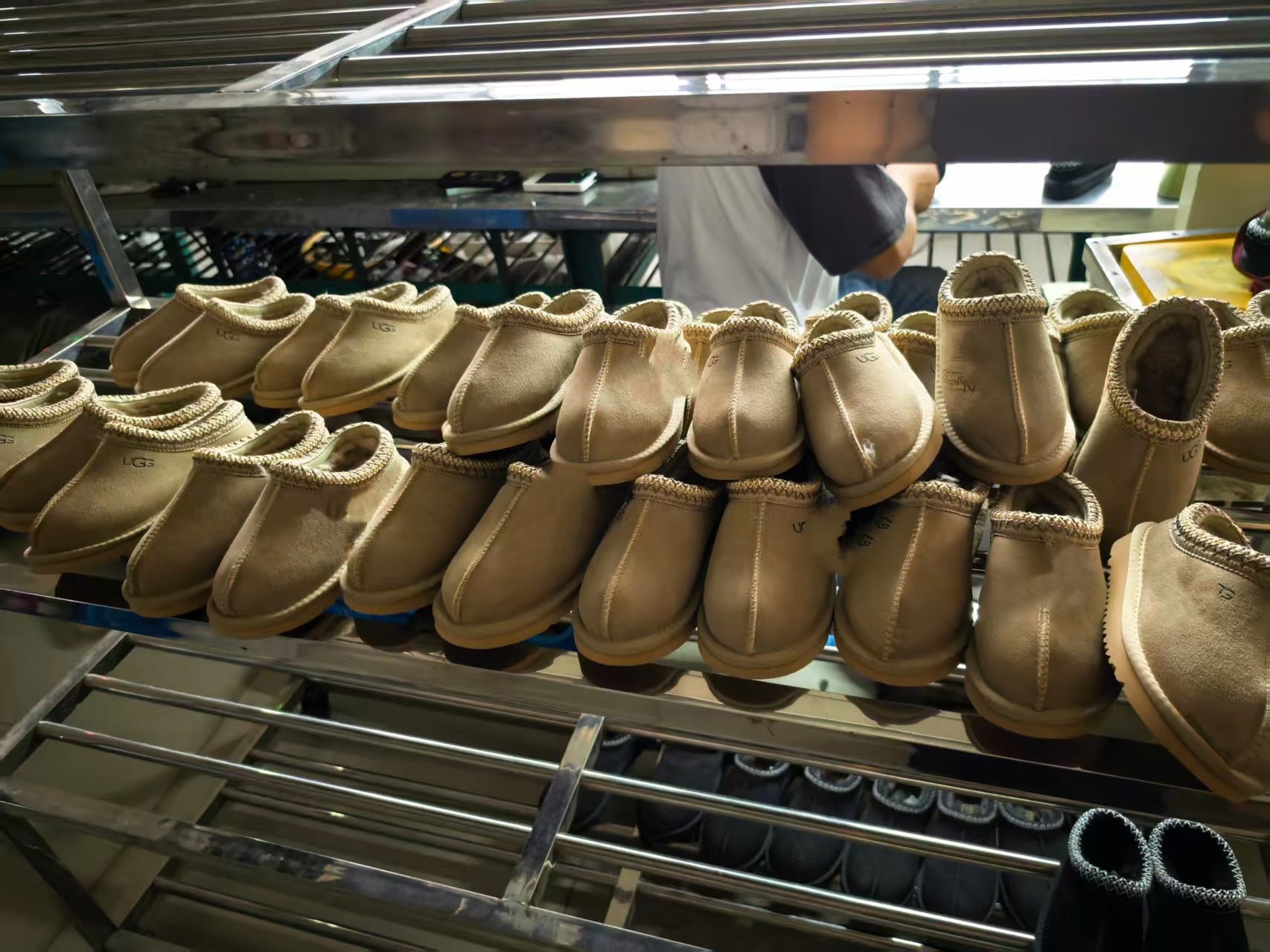 Multiple pairs of tan suede slip-on slippers with white trim displayed on metal racks in a store or storage area.
