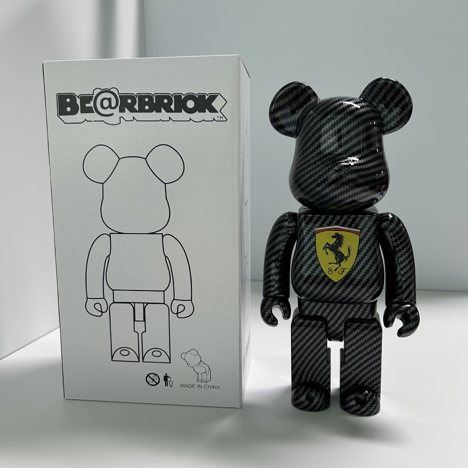 A black carbon-fiber-patterned Bearbrick toy stands next to its white product box, which features a line drawing of the bear figure and a Ferrari shield logo on the chest.