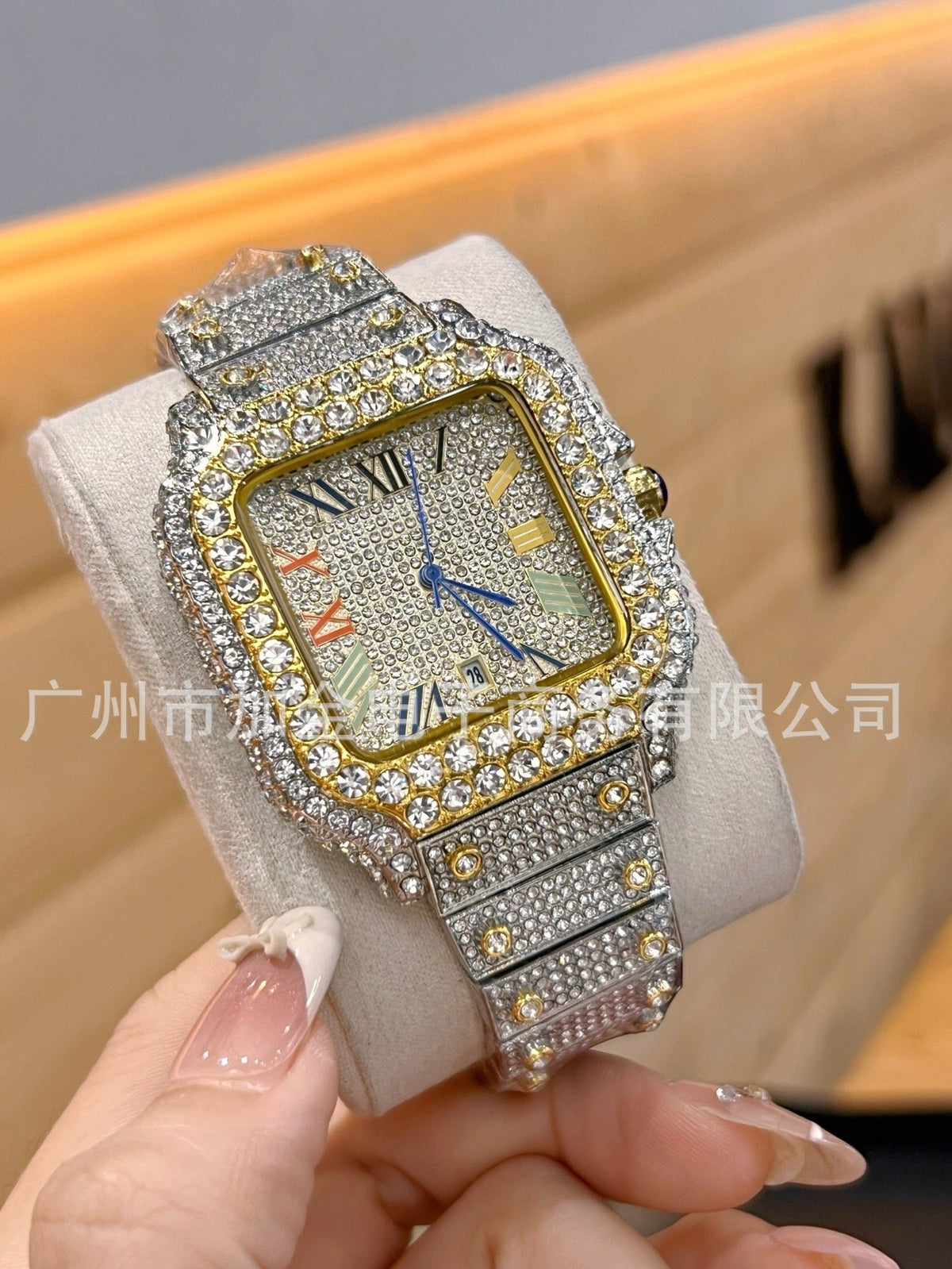 A heavily embellished square wristwatch with a silver-toned metal case and band covered in clear rhinestones, featuring a gold-toned bezel and blue hands, held in a hand against a blurred indoor background.