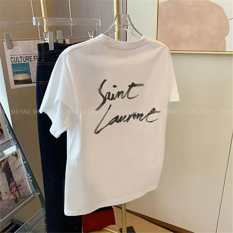 A white short-sleeve T-shirt hanging on a hanger, featuring a large black cursive 'Saint Laurent' logo printed on the back. The shirt is displayed in a retail or studio setting next to dark jeans.
