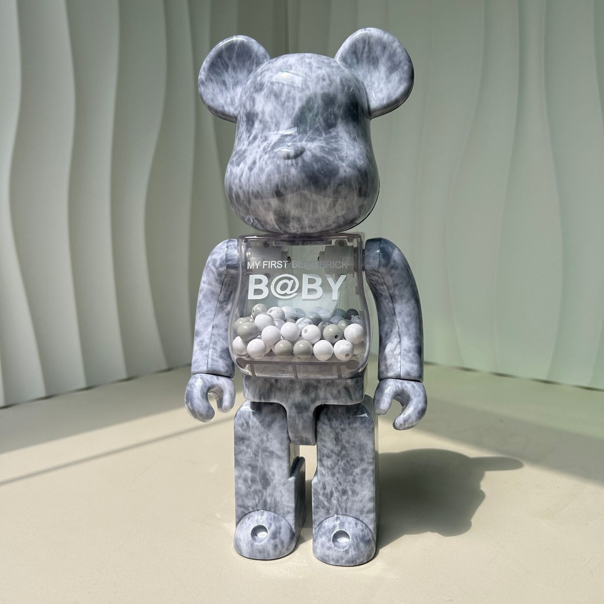 A bear-shaped collectible figure with a mottled gray and white marbled pattern, featuring a transparent chest compartment filled with small white beads and printed with 'MY FIRST B@YY'.