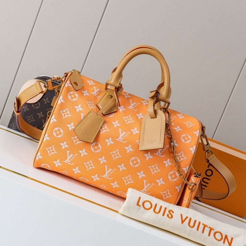 An orange Louis Vuitton Speedy bag with the classic white monogram pattern, featuring tan leather handles and a matching dust bag in the foreground on a white shelf.