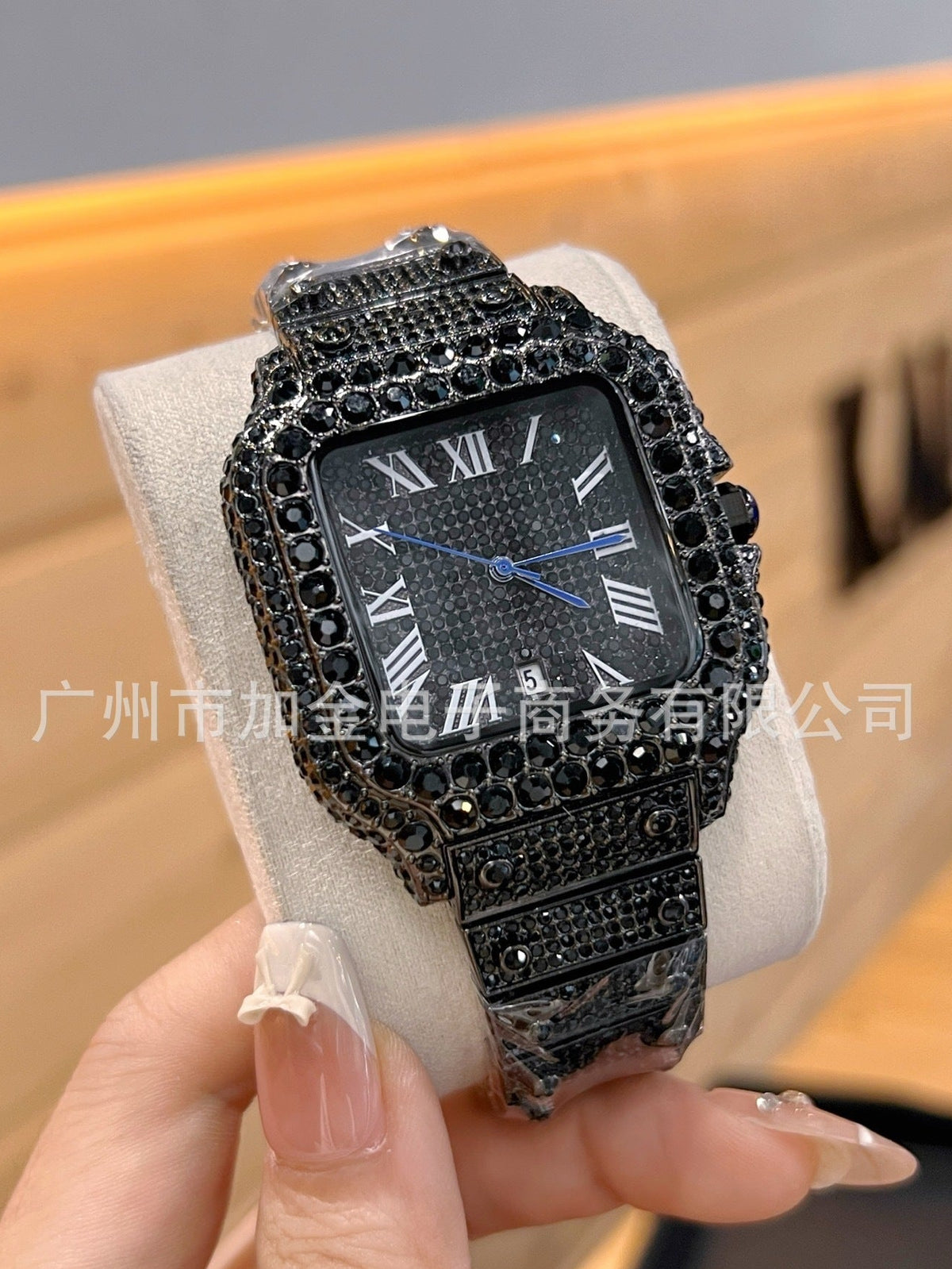 A black watch with a rectangular face, Roman numeral hour markers, and a heavily embellished rhinestone bezel and strap, held in a hand on a display stand.