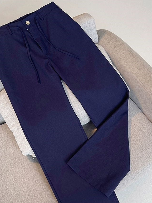 Dark navy blue drawstring trousers laid flat on a light beige fabric sofa. The pants feature a straight-to-slightly-flared leg, a button closure, and a drawstring waistband.