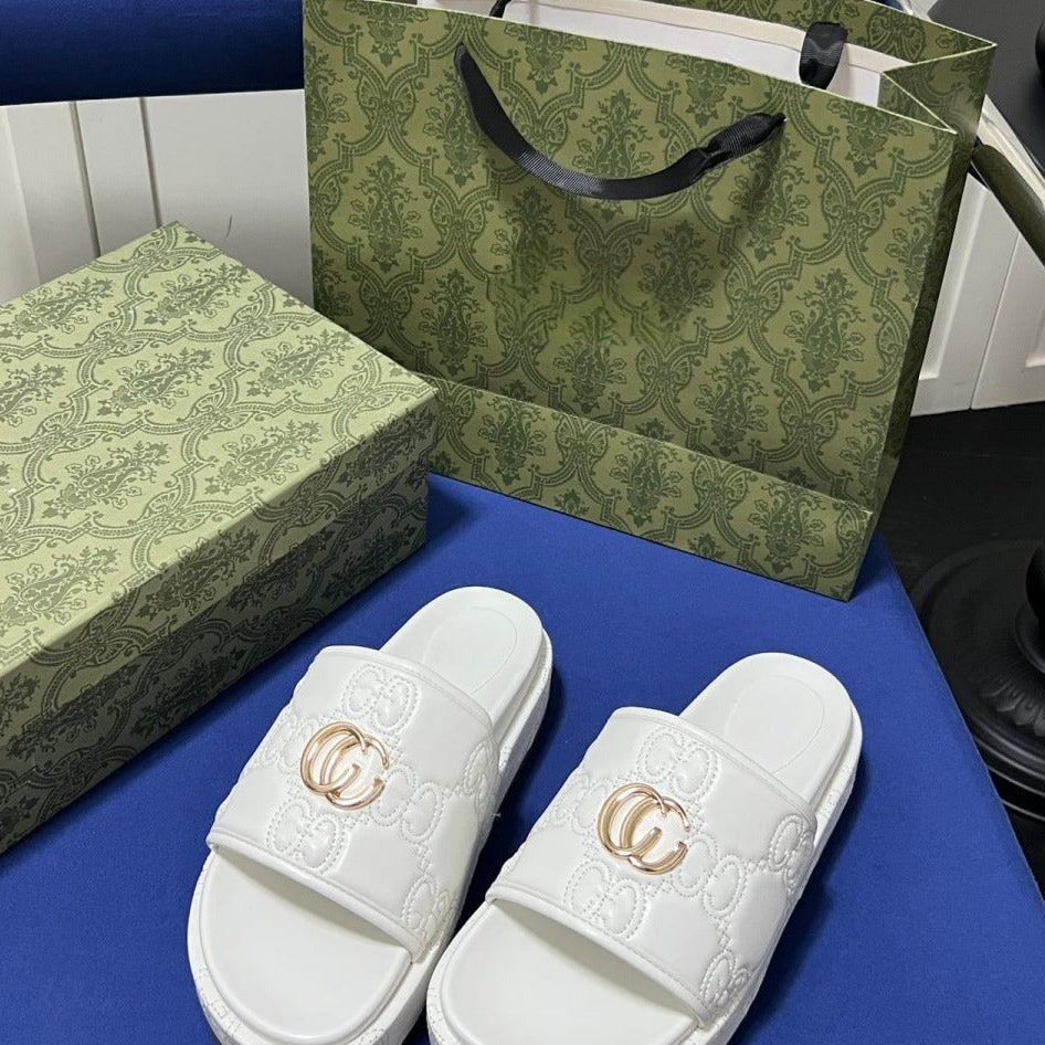 A pair of white Gucci slide sandals with embossed GG monogram patterns and gold-toned double-G hardware, displayed on a blue surface with a matching green patterned box and tote bag behind them.