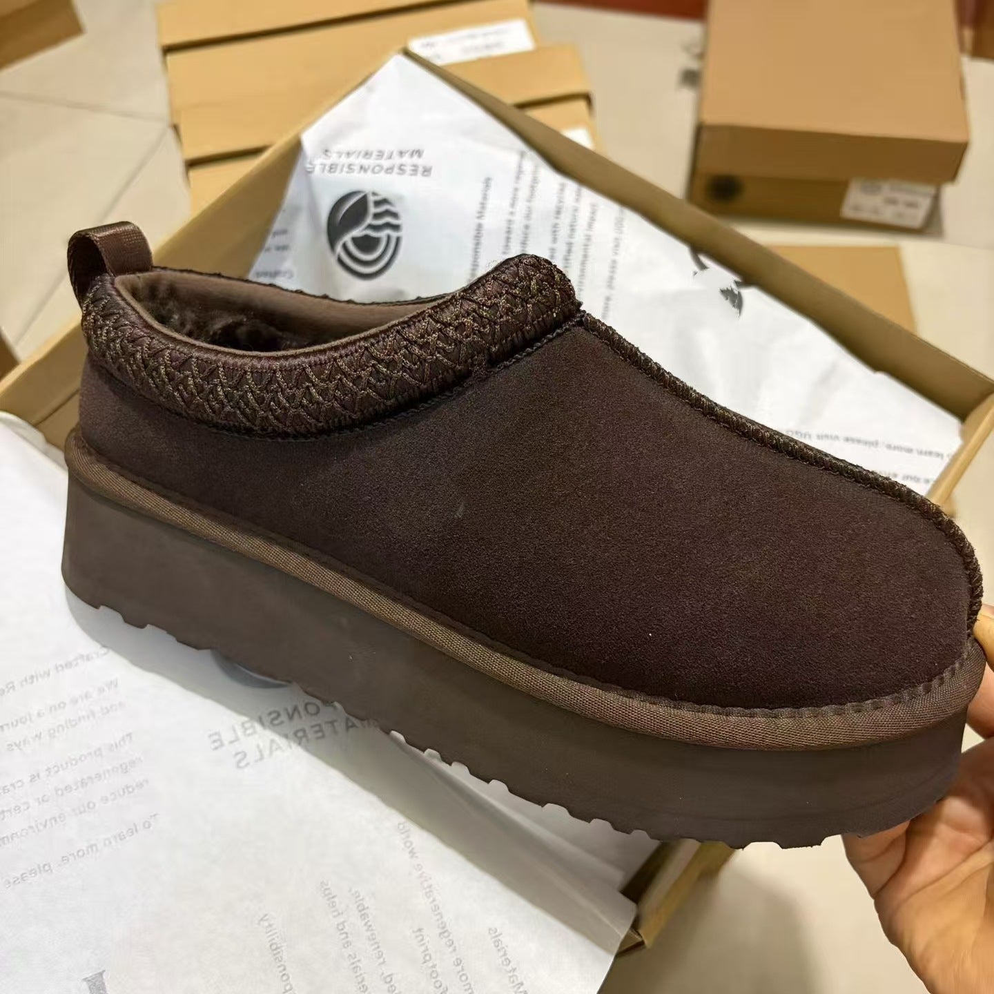 A dark brown slip-on clog-style shoe with a thick platform sole, held over an open shoebox with tissue paper. The shoe has a textured brown knit trim around the collar and a fuzzy brown lining.