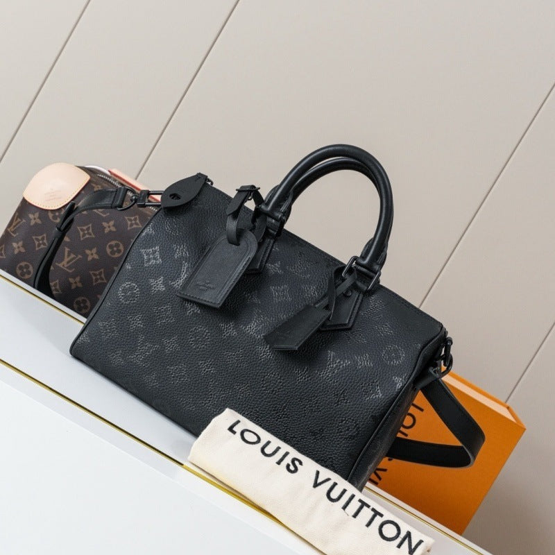 A Louis Vuitton black monogram Speedy handbag displayed on a white shelf next to an orange Louis Vuitton shopping bag and a dust bag with the Louis Vuitton name printed on it.