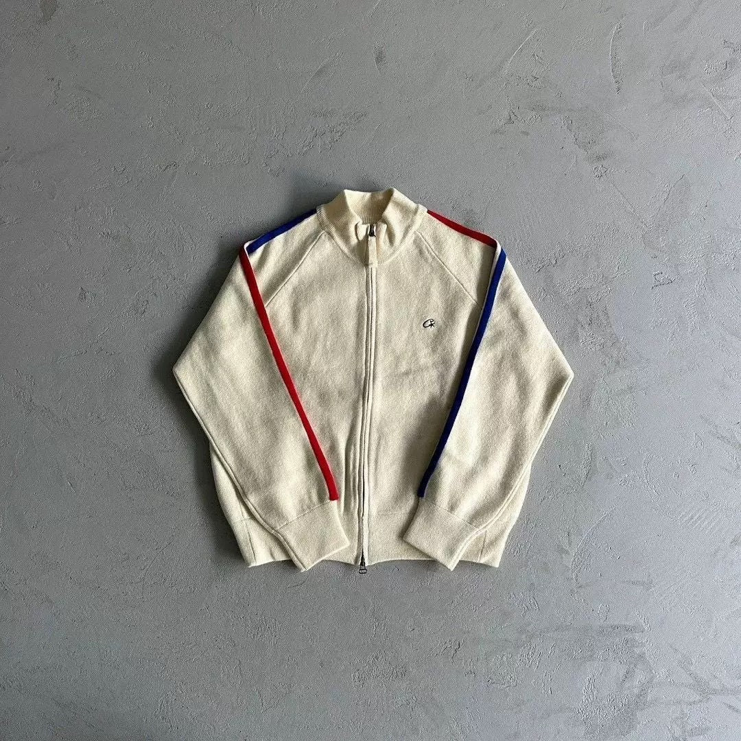 An off-white zip-up track jacket with red and blue shoulder stripes, laid flat on a textured gray background. A Lacoste crocodile logo is visible on the left chest.