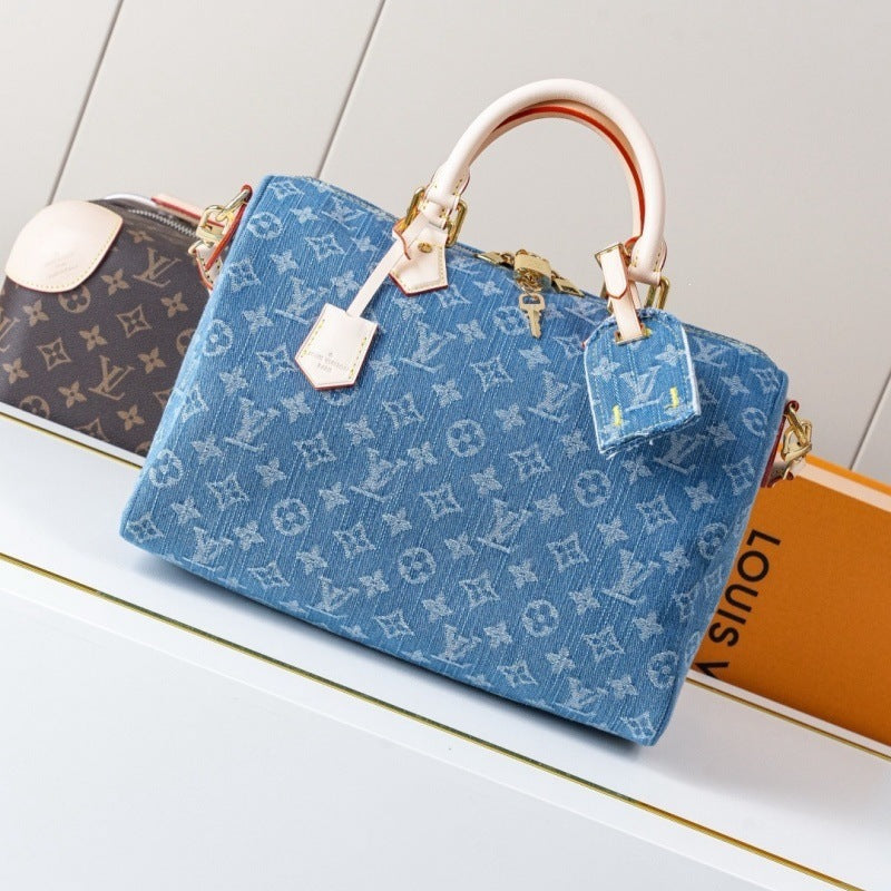 A blue Louis Vuitton monogram canvas handbag with light beige leather top handles and gold-tone hardware, displayed on a white shelf next to other Louis Vuitton items.
