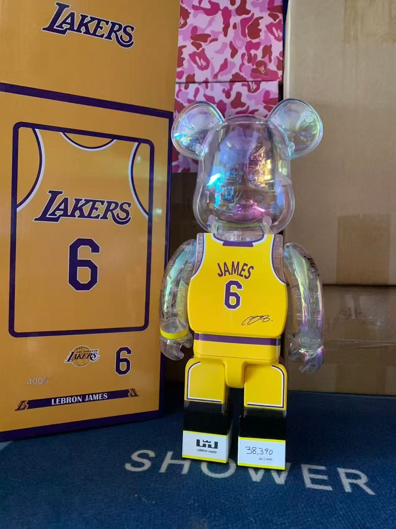 A translucent iridescent bear-shaped collectible figure wearing a yellow and purple Lakers jersey with the number 6, standing in front of its matching Lakers-themed box. The figure has clear plastic ears and limbs with a holographic finish, and the box behind it shows Lakers branding and the number 6.