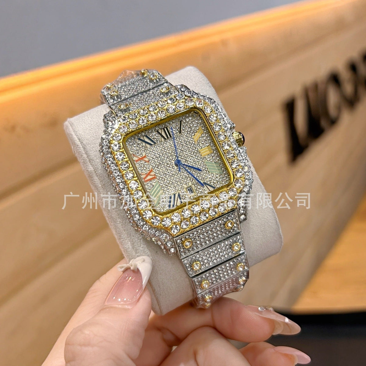 A close-up of a wristwatch with a square gold-tone case and a silver-tone link bracelet, both heavily embellished with clear rhinestones. The watch is being held by a hand with manicured nails, with a wooden display case in the blurred background.
