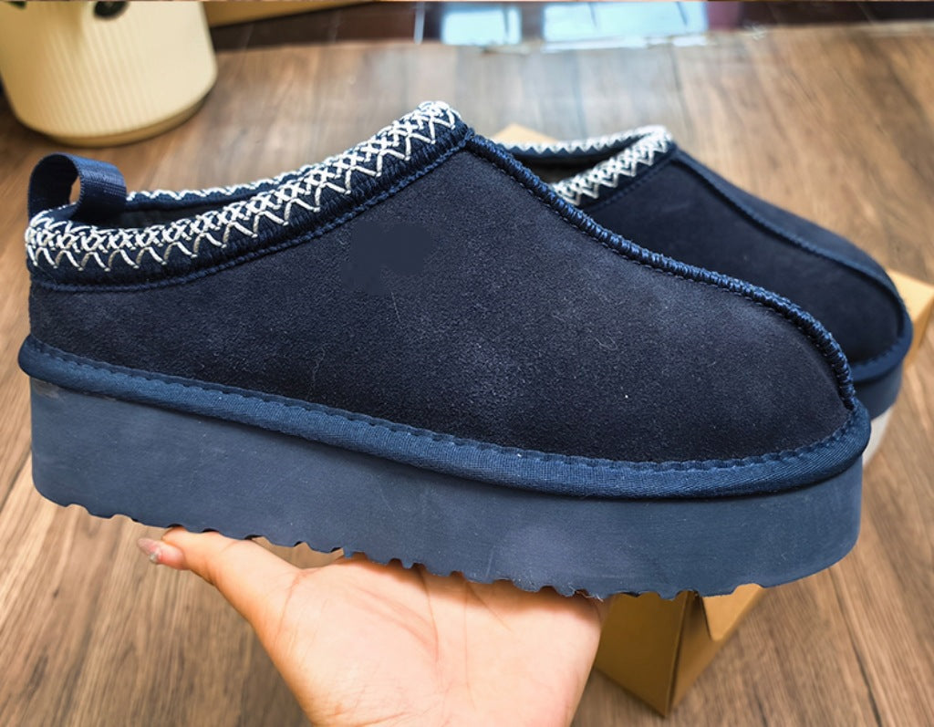A pair of navy blue slip-on mule shoes with white zigzag stitching along the collar, held in a hand over a wooden floor background.