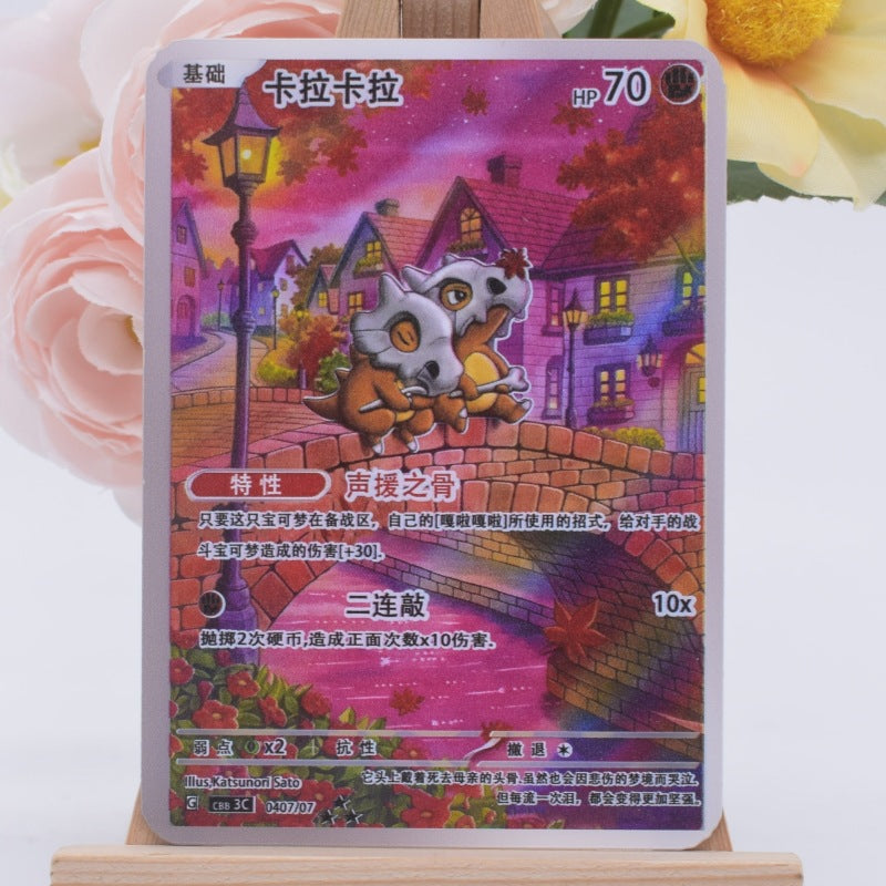 A Pokémon trading card featuring a cute owl-like creature in a colorful village scene, displayed upright on a small wooden stand with soft pink flowers in the background.