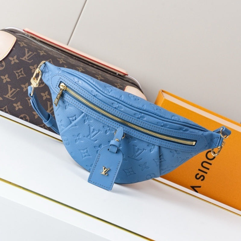 A blue Louis Vuitton embossed leather fanny pack with gold zippers and hardware, displayed on a white surface next to its brown monogram box and an orange box.