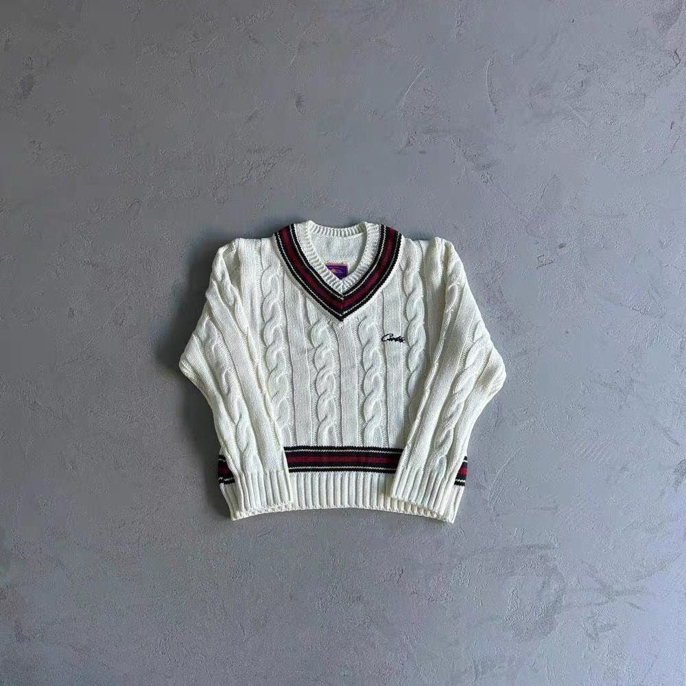 A cream-colored cable-knit sweater with a V-neck and ribbed trim in navy and red, laid flat on a textured gray background.