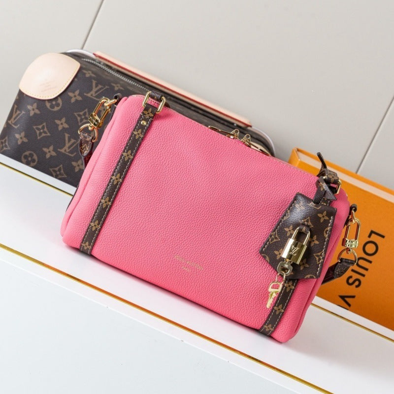 A pink Louis Vuitton pebbled leather shoulder bag with brown monogram canvas straps and a matching lock charm, displayed on a white shelf next to an orange Louis Vuitton box and a monogram pouch.