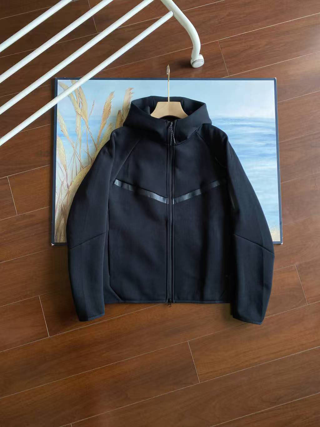 A black hooded zip-up jacket with subtle gray contrast panels on the chest and sleeves, hanging on a wooden hanger against a framed landscape print on a dark wood floor.