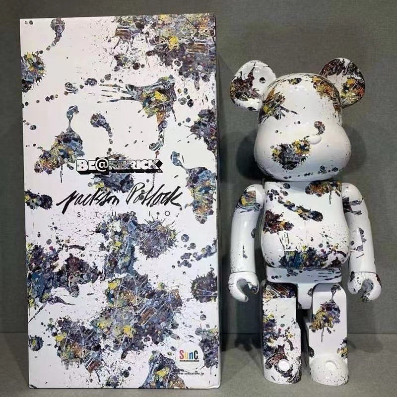 A Bearbrick collectible figure standing next to its retail box. The figure and packaging feature a white base covered in multicolor paint splatters in blue, purple, yellow, and black, with 'BEARBRICK' and 'Jackson Pollock' branding visible.