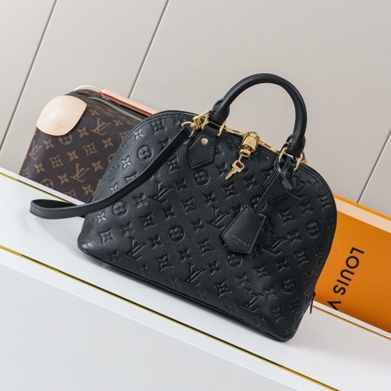 A black Louis Vuitton monogram embossed handbag with gold-tone hardware, displayed on a white surface next to a Louis Vuitton monogram pouch and an orange Louis Vuitton box.
