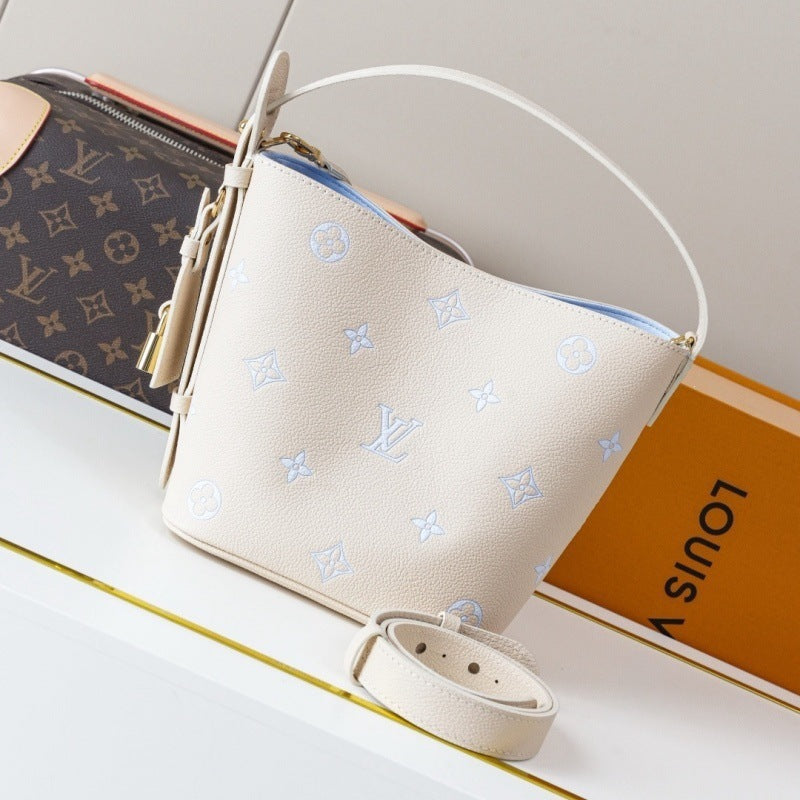 A light beige Louis Vuitton monogram bucket bag with a white top handle and a matching detachable strap, displayed next to its orange and brown monogram packaging on a white surface.