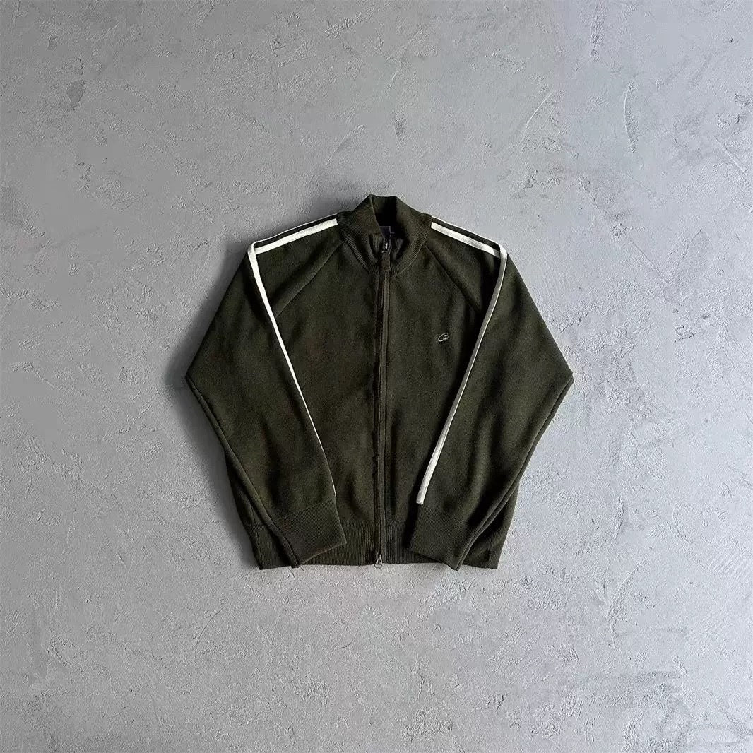 An olive green zip-up track jacket with white stripes on the shoulders, laid flat on a textured gray concrete background.