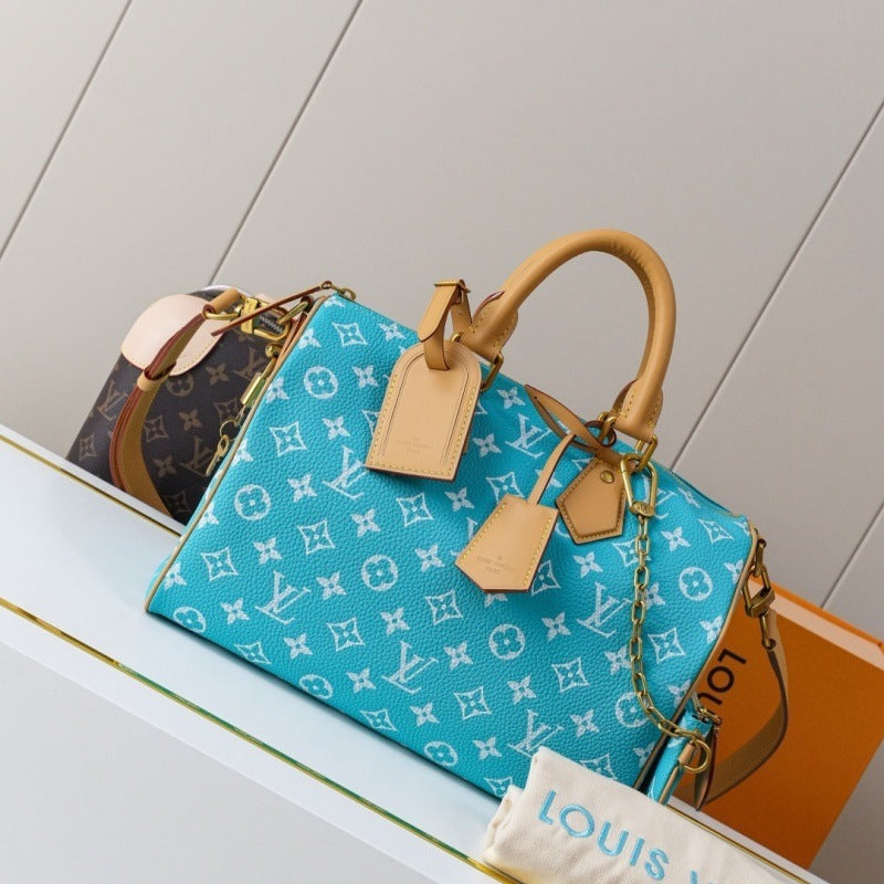 A Louis Vuitton Speedy bag in turquoise blue monogram canvas with tan leather handles and tags, displayed on a white shelf next to another monogram bag and an orange box.